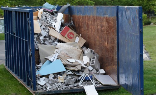 A large, open-top metal skip situated outdoors on a grassy area, filled with assorted waste materials including broken plasterboard, shattered tiles, fragmented wood, and discarded packaging, with some debris piled higher at the front and sides. The skip's exterior shows signs of weathering, with rust spots visible on the inner panel. In the background, there are trees and a clear sky, indicating a rural or suburban setting typical of property clearances in Great Bookham. This scene reflects professional rubbish removal services offered by House Clearance Great Bookham, focusing on the disposal of renovation waste and household debris, suitable for a property cleanout or house clearance in the local area. The overall atmosphere emphasizes operational efficiency in outdoor waste collection, with the waste materials arranged for pickup or disposal, aligning with rubbish removal processes on residential or commercial sites in the town.