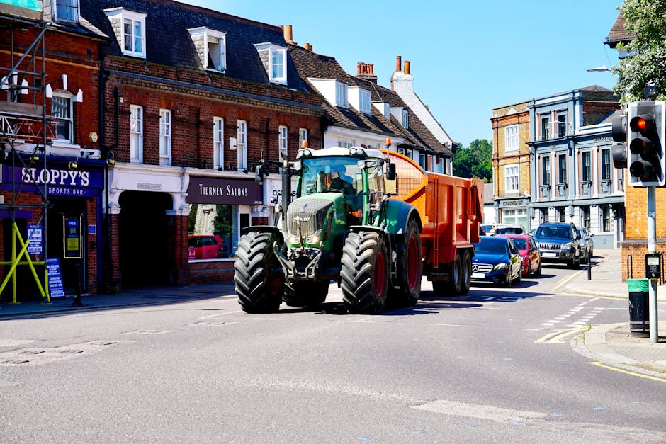 A large green tractor with an orange waste collection container attached to its back is positioned at an urban intersection, partially obstructing the view of several parked cars along the street. The