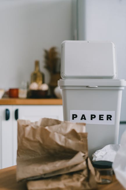 A person in a domestic kitchen, visible from the waist up, is holding a green plastic bottle with both hands, preparing to dispose of it. The individual is dressed in a white t-shirt and has a burnt o