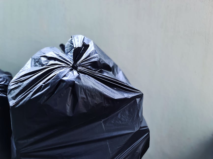 A collection of overflowing rubbish bins and discarded waste bags piled on a paved sidewalk in front of a commercial building. The waste includes cardboard boxes, plastic bags, and paper packaging, so