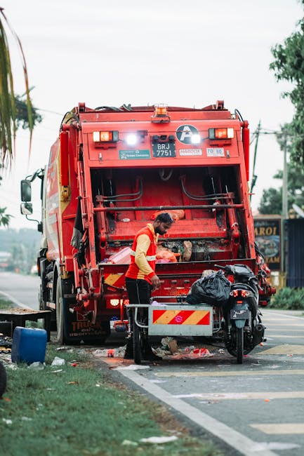 A worker wearing a high-visibility orange vest and dark trousers is standing behind a red refuse collection truck, which is positioned on the side of a street. The truck's rear loading mechanism is op